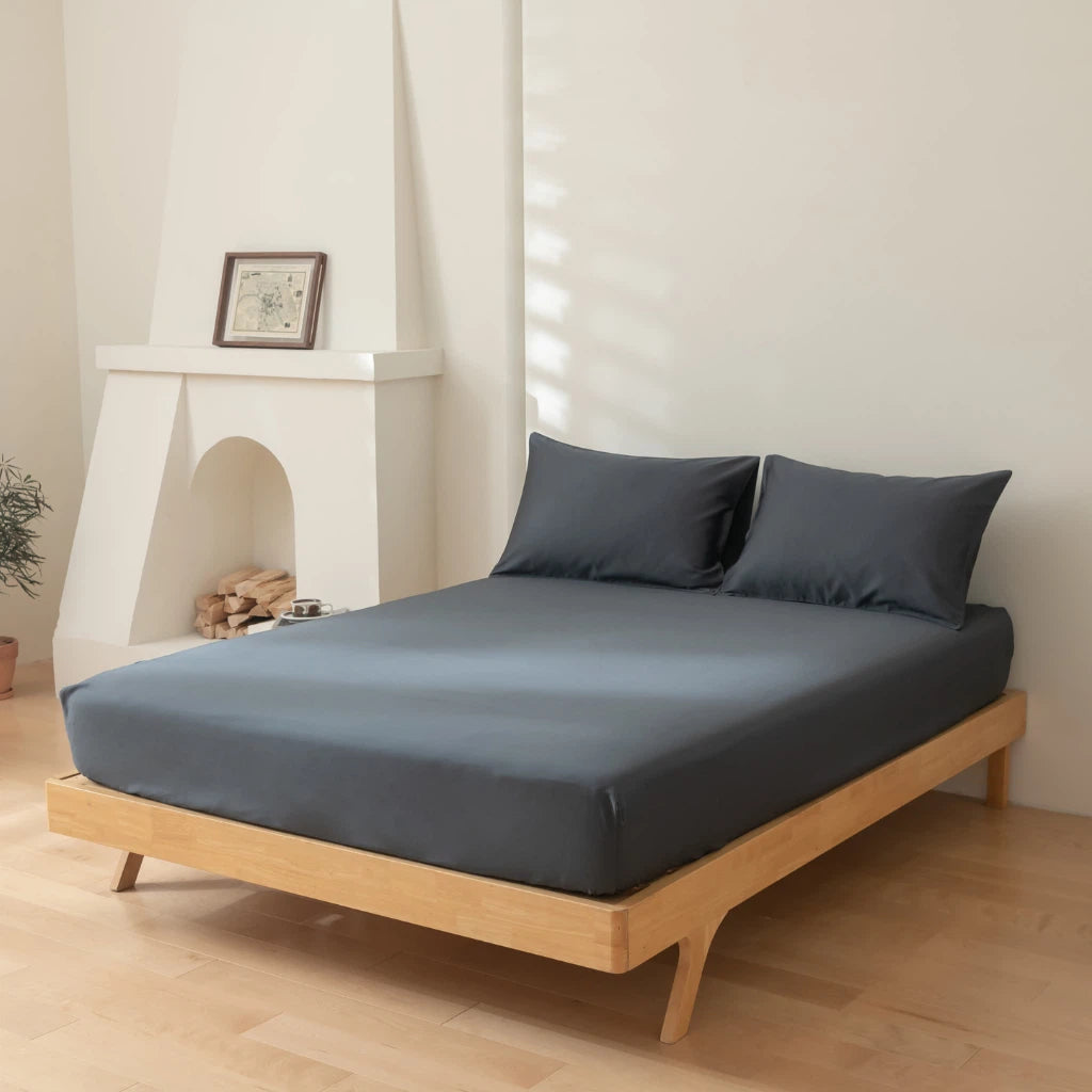 A neatly made bed with a dark grey sheet set in a room with a minimalist design, hardwood floors, and a white fireplace.