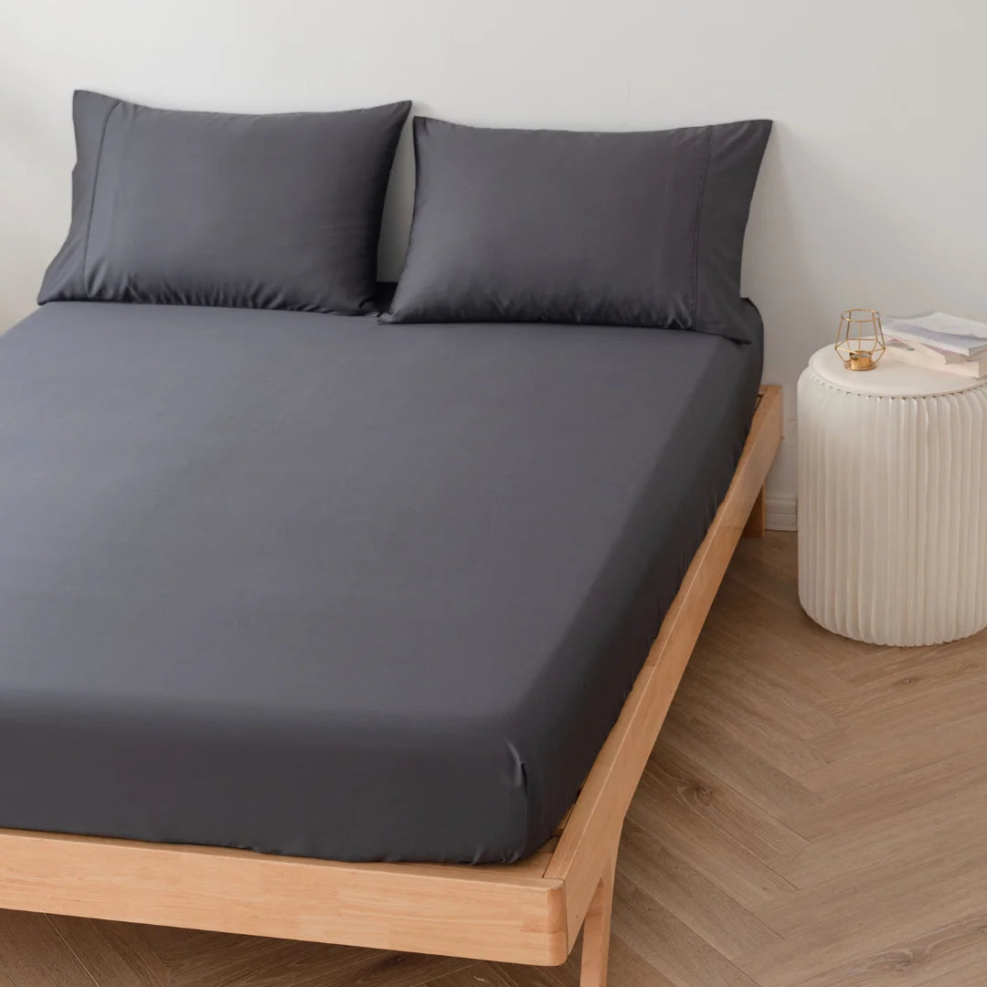 A neatly made bed with Linenly's Bamboo Fitted Sheet in Charcoal and pillowcases on a minimalist wooden bed frame, next to a simple white bedside table.