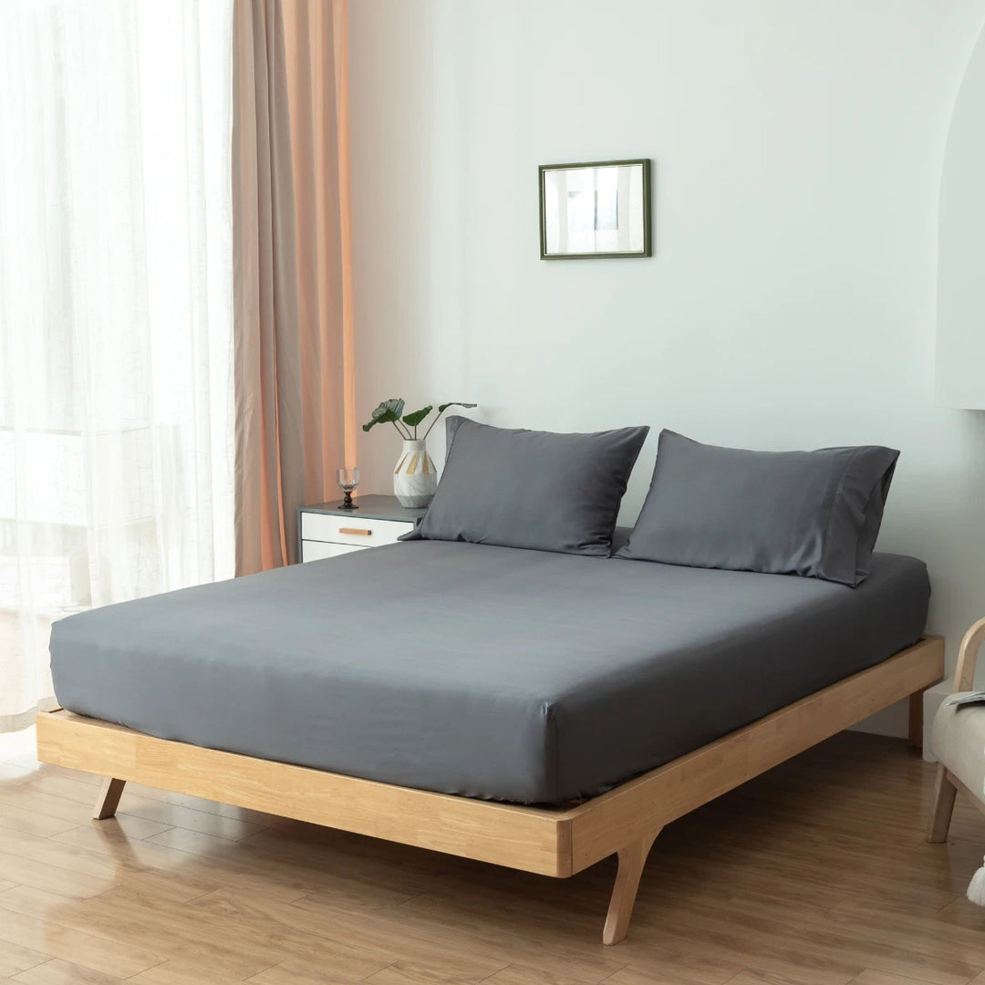 A neatly made bed with Linenly's granite grey bamboo fitted sheets showcasing sumptuous softness in a minimalist bedroom with white walls, a wooden bed frame, a small bedside table, and light curtains.