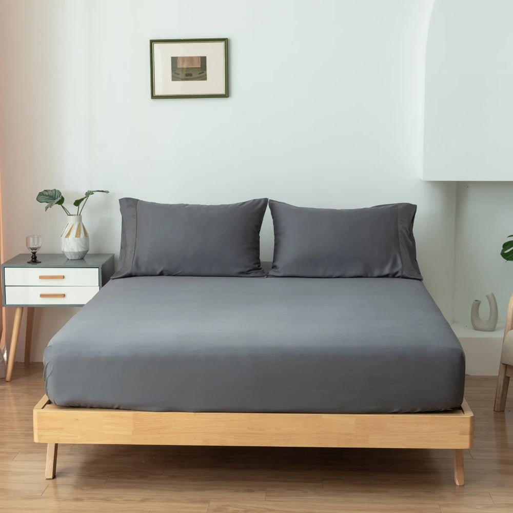 A neatly made bed with Linenly's Granite Grey Bamboo Fitted Sheets in a minimalist bedroom with a white nightstand and decorative vase on the side, offering sumptuous softness and a deep-pocket design.