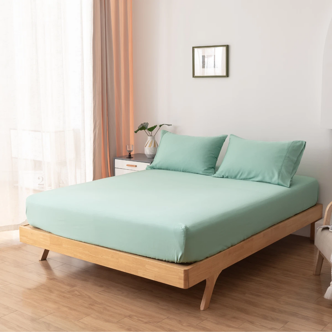 A modern bedroom with a wooden bed, Linenly Bamboo Fitted Sheet - Summer Green, light wood floors, a small bedside table with decor, light curtains, and a framed picture on the white wall.