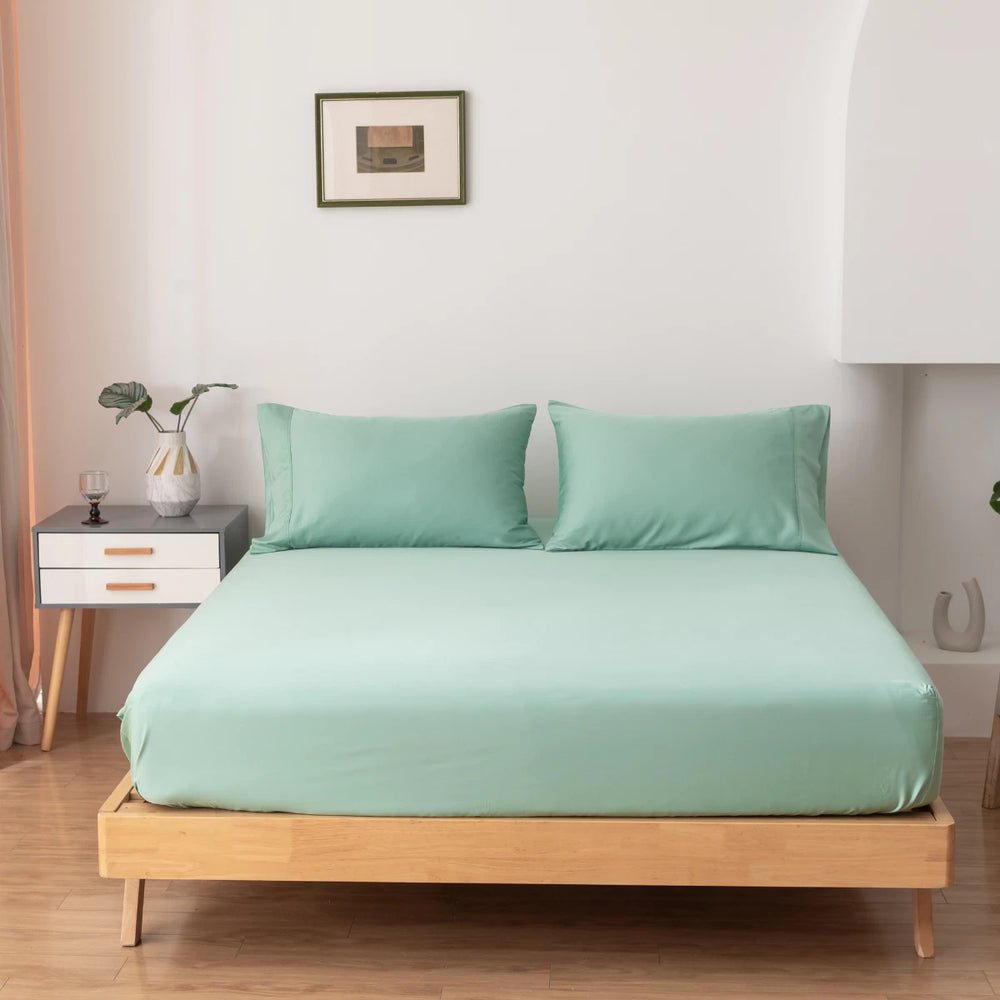 A neatly made bed features Linenly's Bamboo Fitted Sheet in Summer Green and matching pillows on a wooden frame. A nightstand holds a plant, glass, and book, with a framed picture hanging above on the white wall.