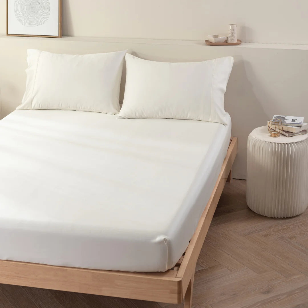 A neatly made bed with crisp Linenly Ivory Bamboo Fitted Sheets on a simple wooden frame, accompanied by a round bedside table with books and a candle, in a serene minimalist bedroom setting.