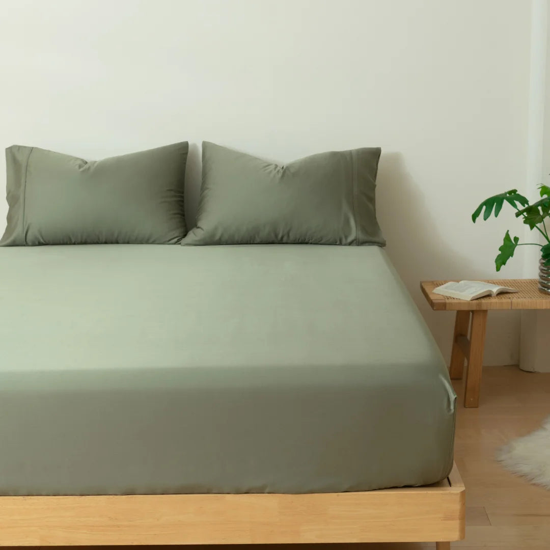 A neatly made bed with Linenly's Bamboo Fitted Sheet in Moss in a minimalist bedroom, featuring a wooden bed frame, a small bedside table with a book, and a potted plant.