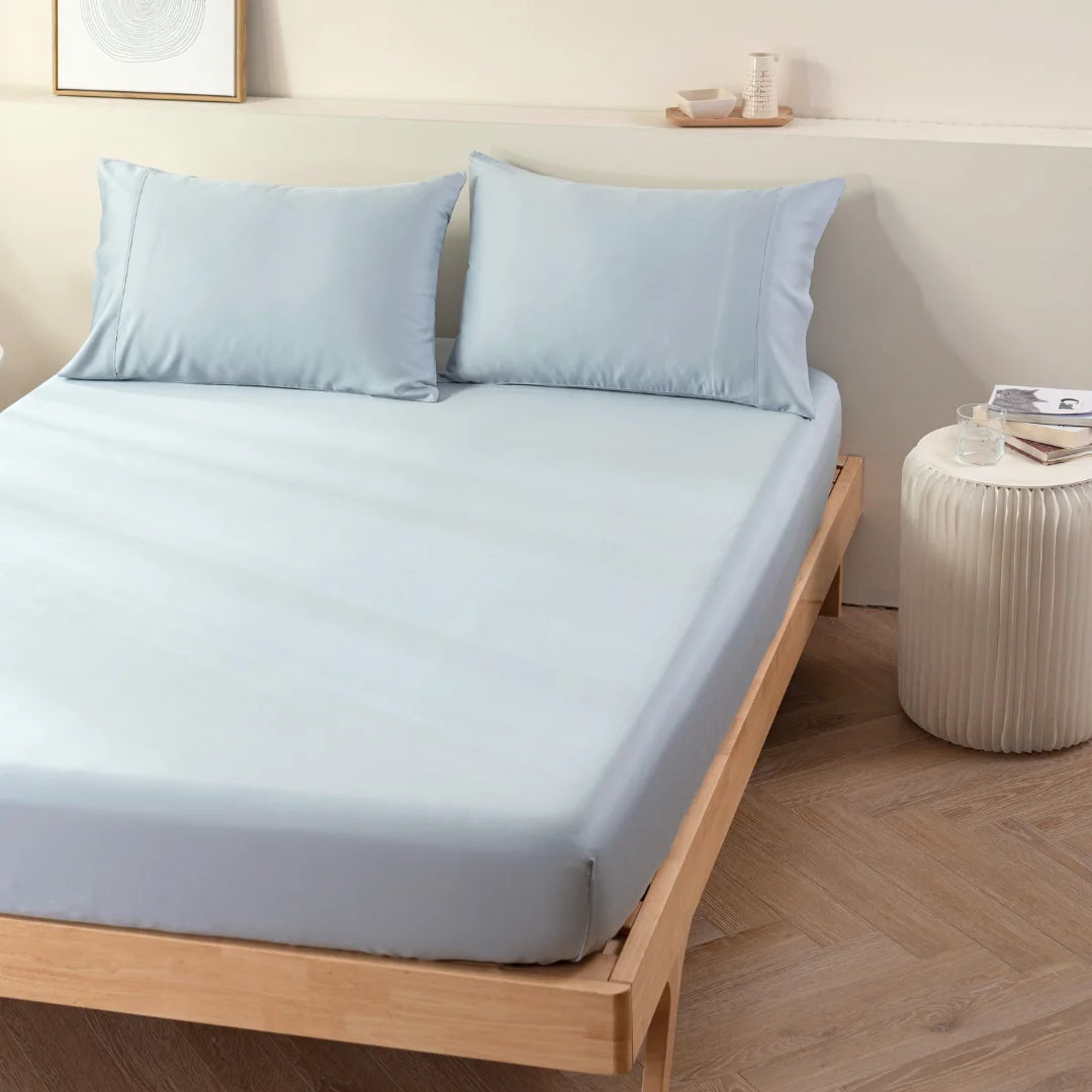 A neatly made bed with Linenly's Bamboo Fitted Sheet in Pale Blue on a simple wooden frame, complemented by a minimalist nightstand and calming wall art in a serene bedroom setting.