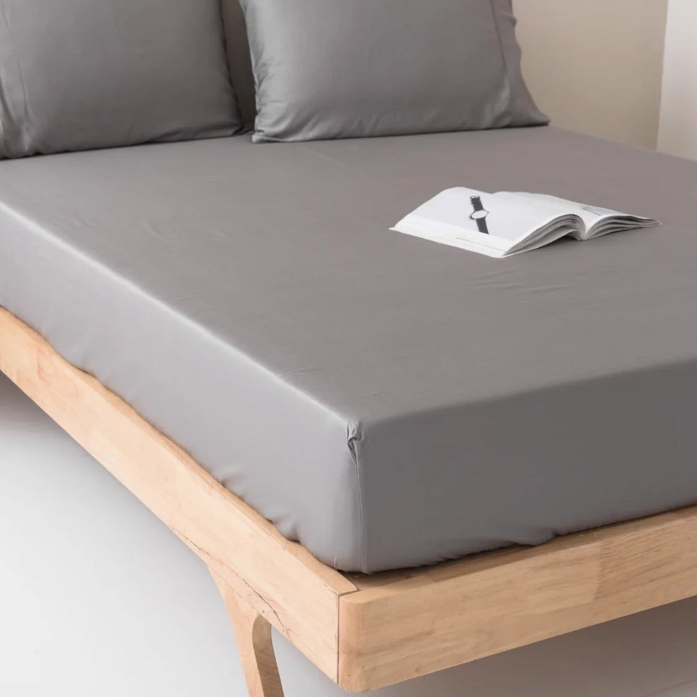 A neatly made bed with a light wooden frame, dressed with luxuriously soft, Linenly stone grey bamboo fitted sheets, and an open book placed in the center, suggesting a quiet moment for reading and relaxation.