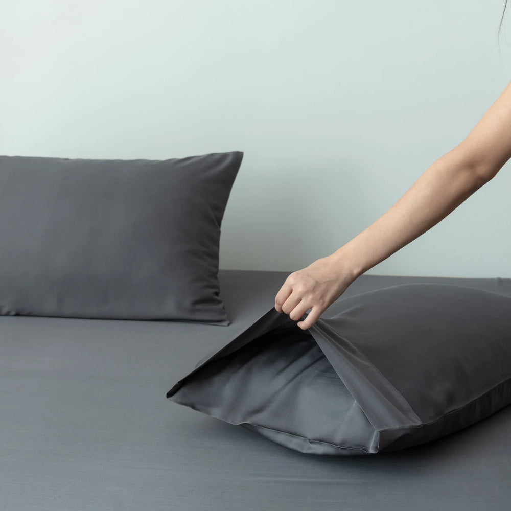 A person's hand gently arranging a dark gray pillow enveloped in Linenly Granite Grey bamboo pillowcases on a neatly made bed with matching sheets in a serene bedroom setting.