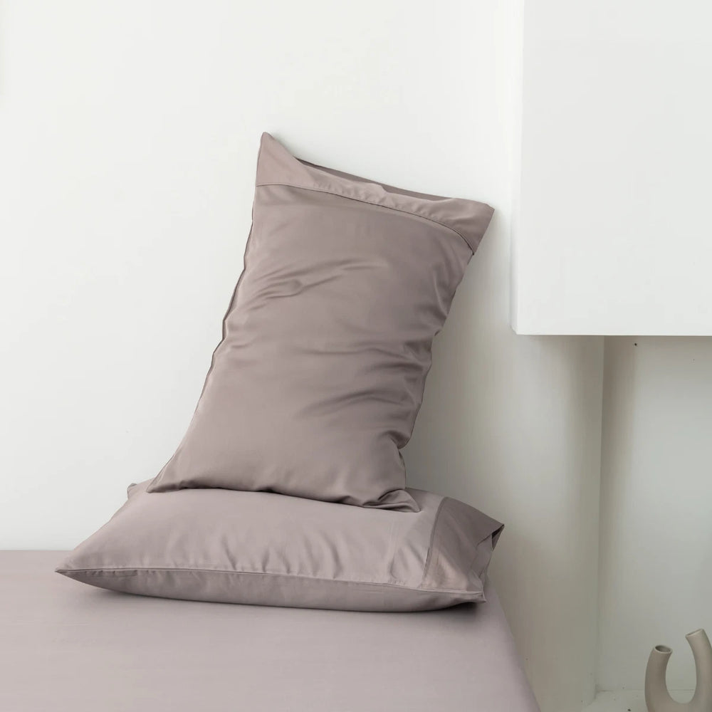 A taupe pillow, encased in a Linenly Bamboo Pillowcase Set - Stone Terrace with a satin weave, casually propped against the wall on a matching bedsheet, next to a white bedside lamp.