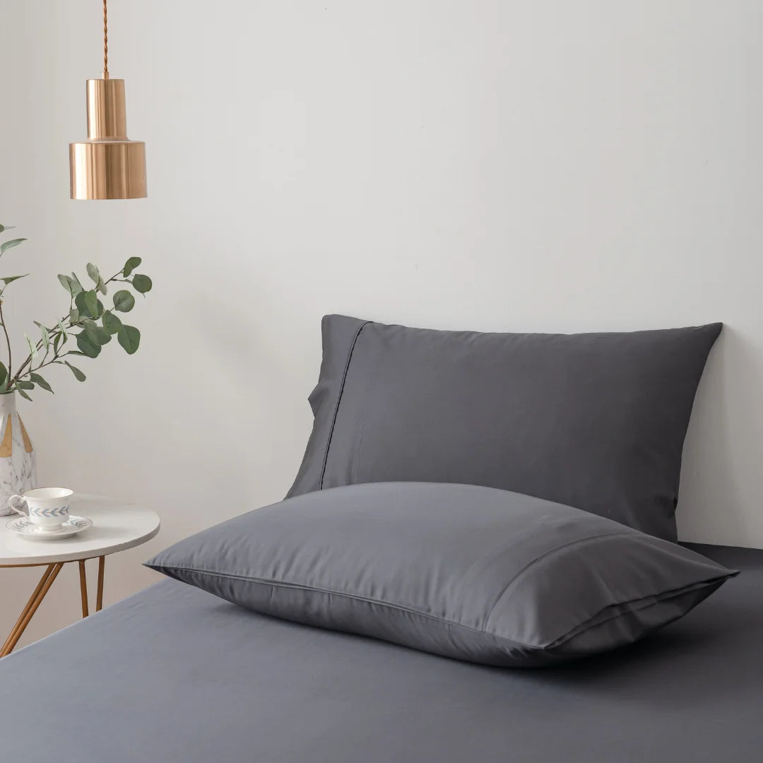 A minimalist bedroom setting with a neatly made bed, a single copper pendant light, an organic Linenly bamboo pillowcase set in Charcoal propped against the headboard, an extra pillow resting on the bed, and a side table adorned.