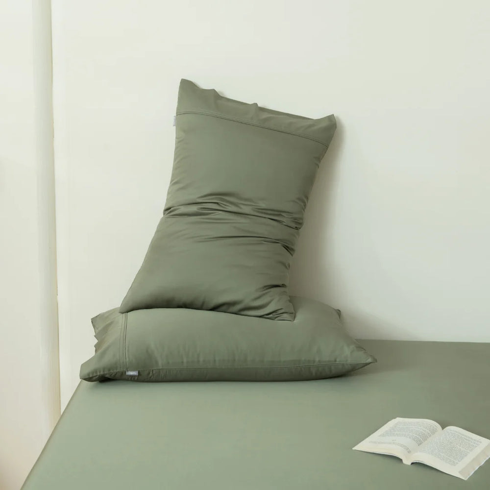 A cozy reading nook with an olive green pillow, featuring Linenly's Bamboo Pillowcase Set in Moss for a cooling effect, stacked against a wall and an open book waiting to be read on a simple cushioned surface.