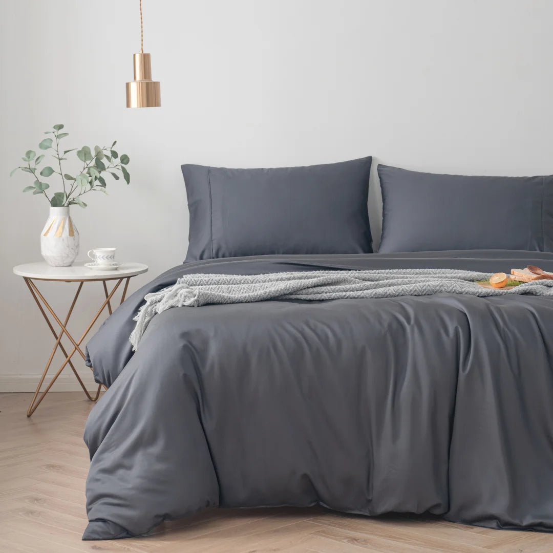 Modern bedroom with a neatly made bed featuring an eco-friendly Linenly bamboo quilt cover in charcoal, complemented by a small side table with a plant and a cozy throw blanket, creating a tranquil and minimalist atmosphere.
