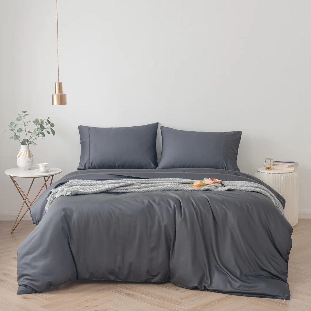 A neatly made bed with a Linenly eco-friendly Charcoal Bamboo Quilt Cover in a minimalist bedroom with a white side table, a small plant, and a hanging light fixture.