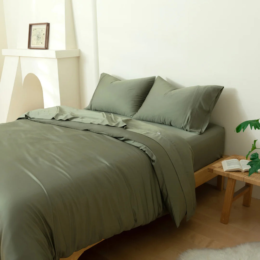 A neatly-made bed with a Linenly bamboo quilt cover in moss color in a minimalist bedroom with a wooden side table, a plant, and a book, creating a serene and comfortable atmosphere.