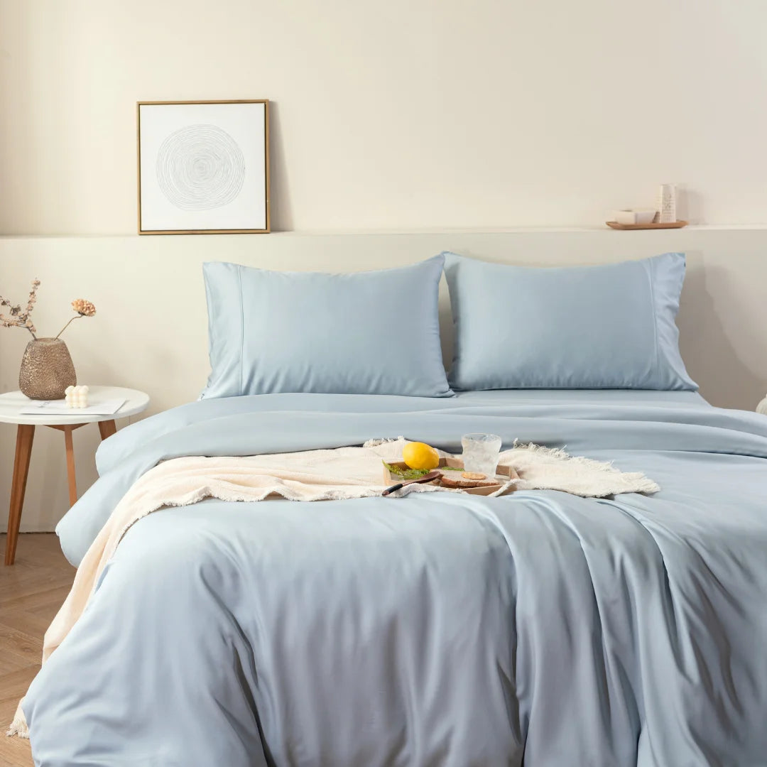 A neatly made bed with a Linenly pale blue bamboo quilt cover in a tranquil bedroom setting, adorned with a breakfast tray and a touch of decorative flora.