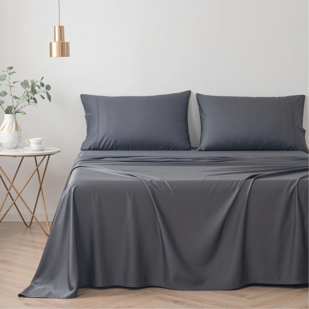 A neatly made bed with smooth Linenly Charcoal Bamboo Sheet Set in a serene bedroom with minimalist decor, featuring a bedside table with an eco-friendly plant and a simple pendant light.