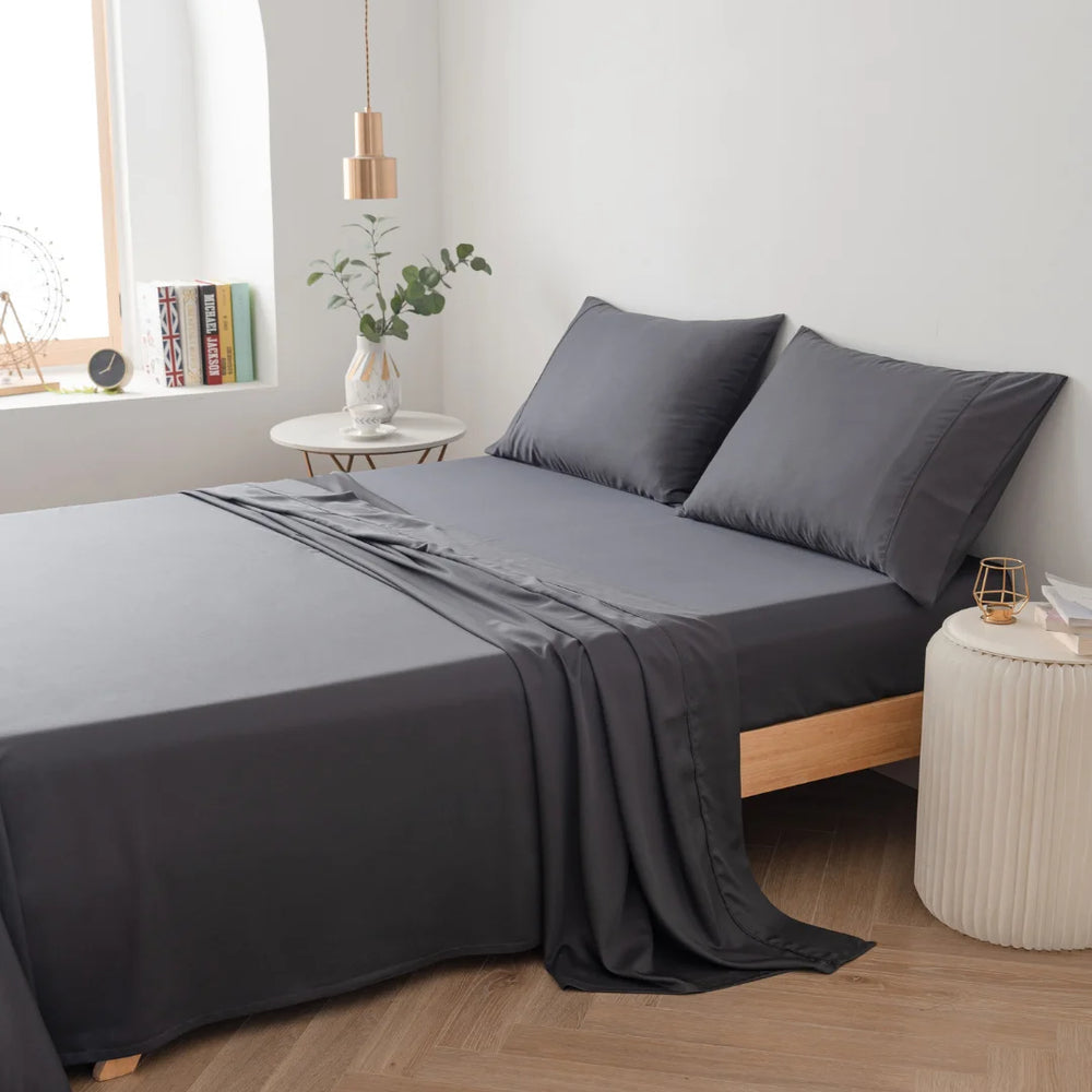 Modern minimalist bedroom with neatly-made bed featuring luxurious Linenly charcoal bamboo sheet set, beside a small round side table with a plant, on a clean and simple backdrop.