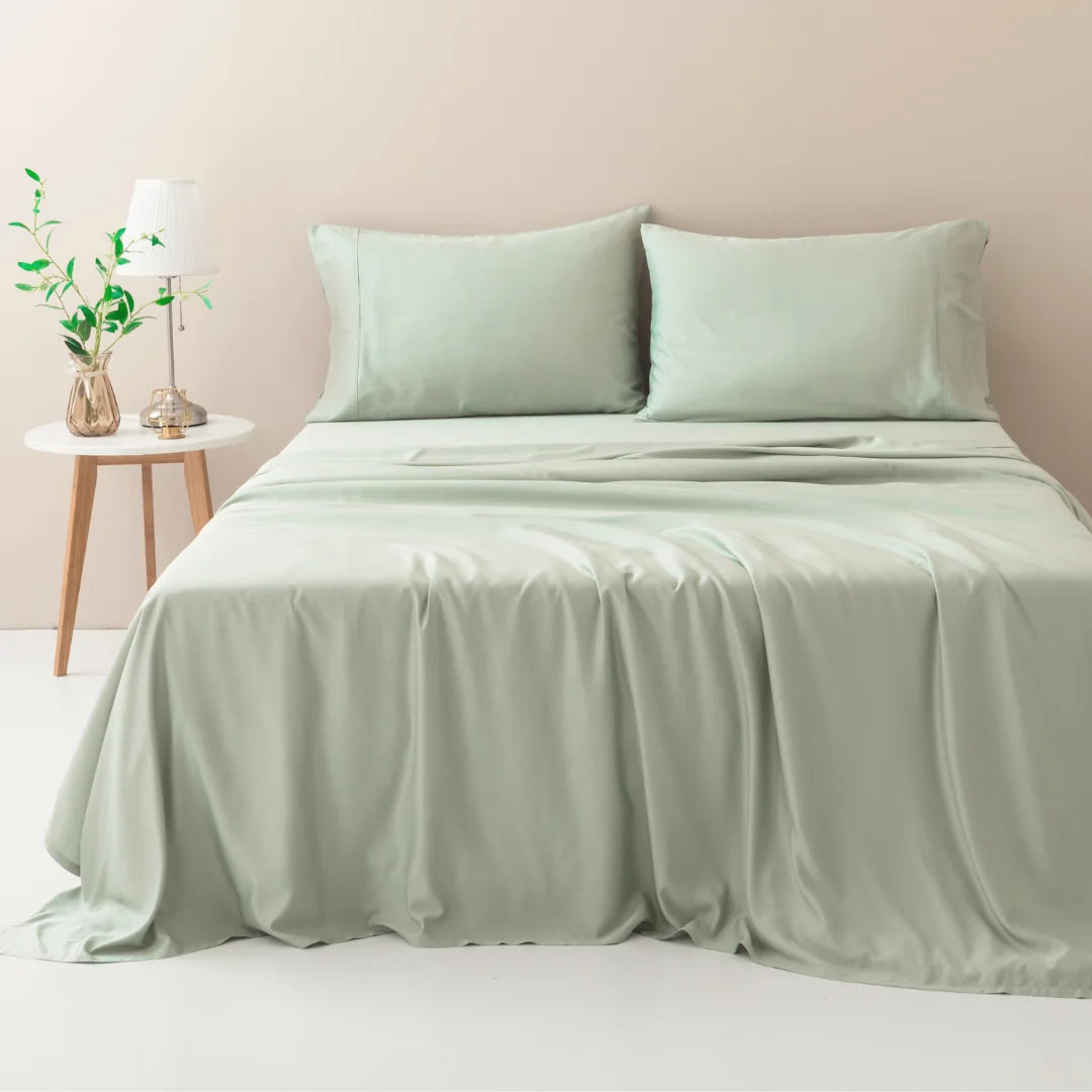 A neatly made bed with luxuriously soft Linenly Sage Bamboo Sheet Set in a minimalist bedroom, accompanied by a small side table with a lamp and a vase of fresh greenery.