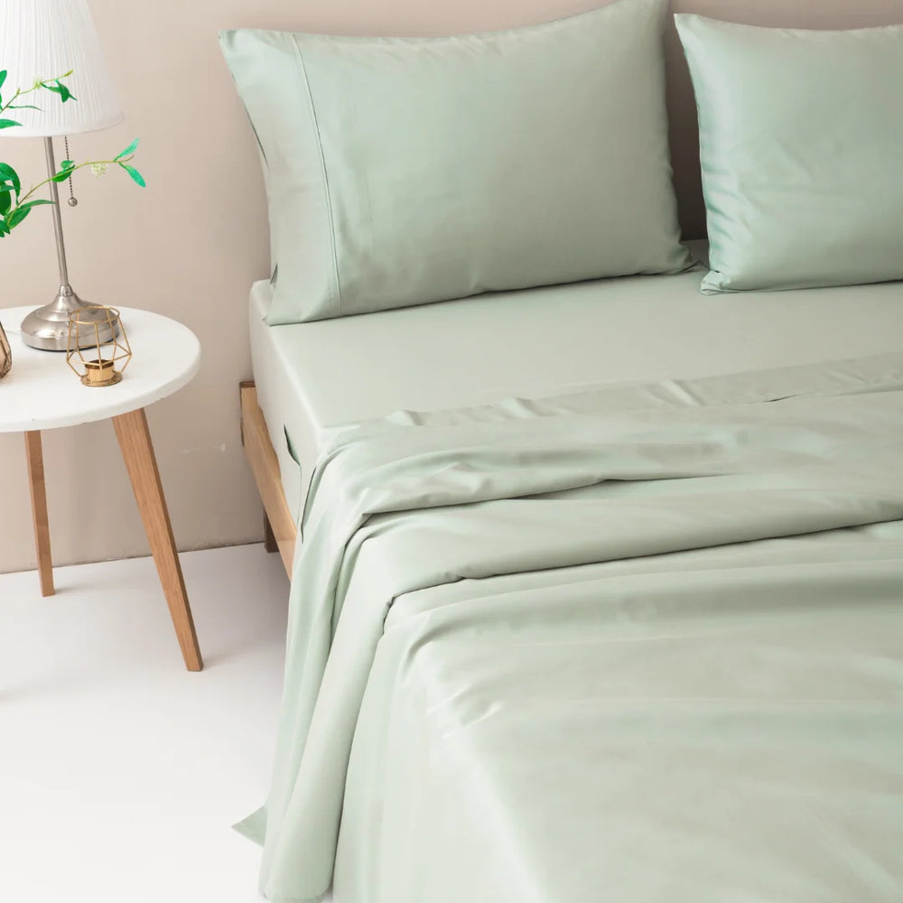A neatly made bed with luxuriously soft Linenly Sage Bamboo Sheet Set in a tranquil bedroom setting, accented by a small wooden bedside table and a vase with fresh greenery.