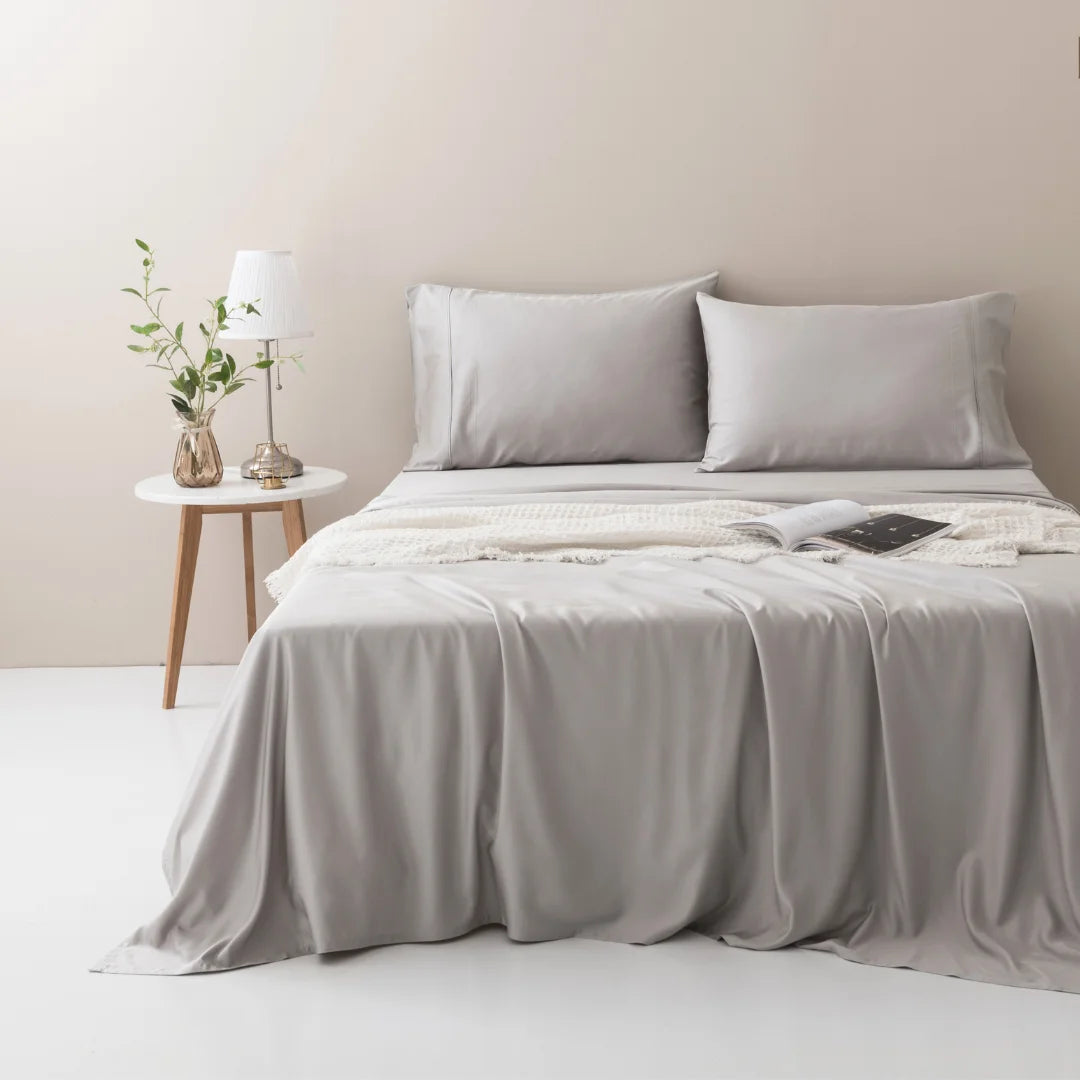 A neatly made bed with a luxury, cooling Linenly Silver Bamboo Sheet Set, complemented by a wooden bedside table with a lamp and a vase of greenery, in a soothing minimalist bedroom setting.