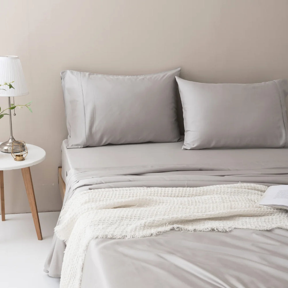 A neatly made bed with Linenly's luxurious, cooling silver bamboo sheet set and a cozy cream blanket, accompanied by a small side table with a lamp and a book, creating a serene and inviting bedroom atmosphere.