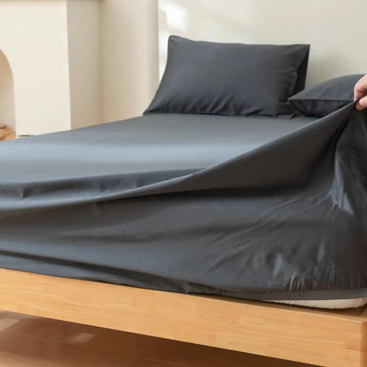 A hand pulls back a Linenly Luxe Sateen Charcoal fitted sheet, crafted from long-staple cotton, to reveal the mattress on a neatly made wooden bed.
