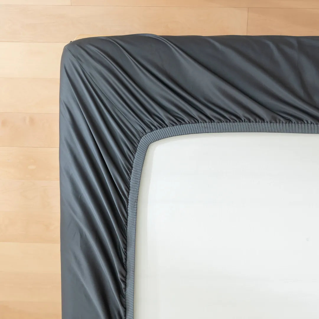 A Linenly Luxe Sateen Fitted Sheet in Charcoal neatly hugging the corner of a mattress on a wooden bed frame, epitomizing modern elegance.