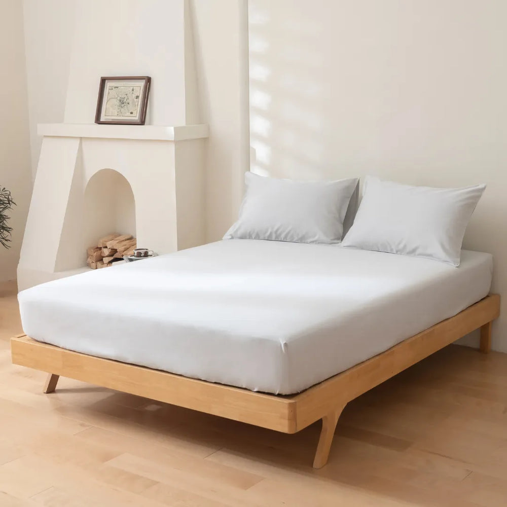 A neatly made bed with a Linenly Luxe Sateen Fitted Sheet in Silver on a modern wooden frame, situated in a minimalist bedroom with a decorative fireplace and a touch of greenery.