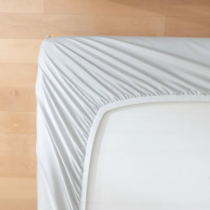 A neatly made bed with a Linenly Luxe Sateen Fitted Sheet in Silver on a wooden floor, exhibiting a minimalistic and tidy bedroom setting.