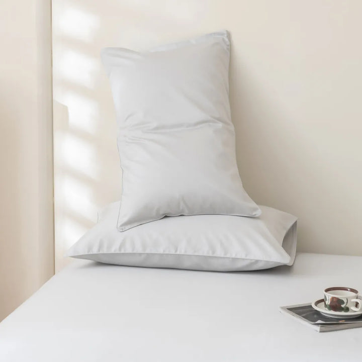 A single white pillow, crafted from premium long staple cotton with a sateen weave, resting against a soft-toned wall with sunlight filtering through blinds, accompanied by a neatly placed Linenly Luxe Sateen Flat Sheet in Silver.
