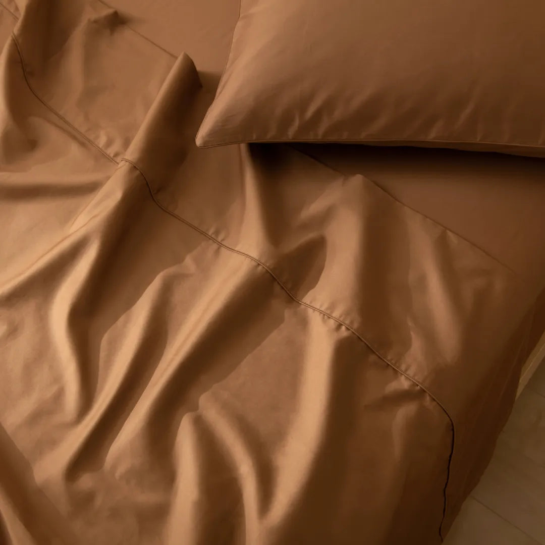 A close-up of a neatly made bed with a Luxe Sateen Flat Sheet in Terracotta from Linenly, along with smooth, premium long staple cotton bedding in earth tones and matching pillows suggesting a warm and cozy atmosphere.