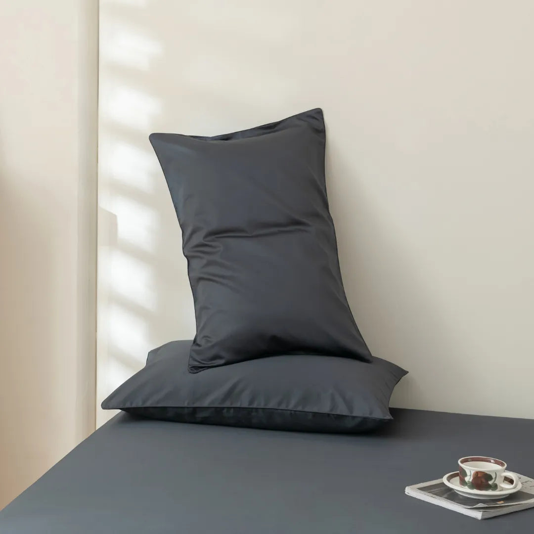 A minimalist setting with a neatly arranged dark pillow, enveloped in Linenly's Luxe Sateen Pillowcase Set in Charcoal, on top of another against a pale wall, beside a small tray with a cup and saucer.