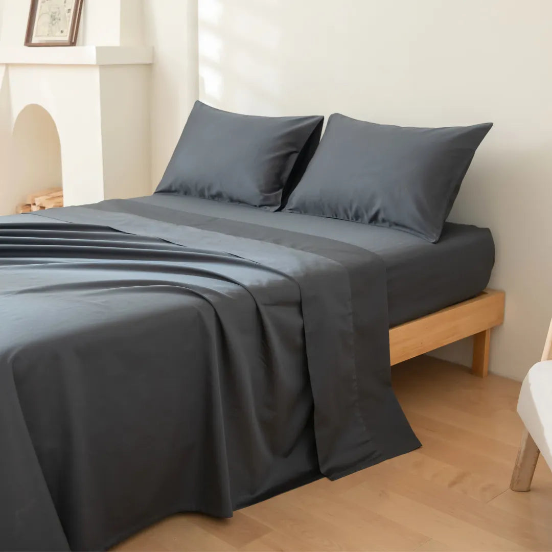 A neatly made bed with a wooden frame, dressed in smooth gray Linenly Luxe Sateen Pillowcase Set - Charcoal bedding, in a room with light walls and minimal decor.