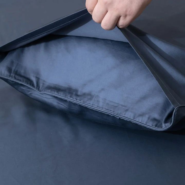 A hand flipping over a Luxe Sateen Pillowcase Set - Midnight by Linenly with an envelope closure design to show its inside against a matching blue background.