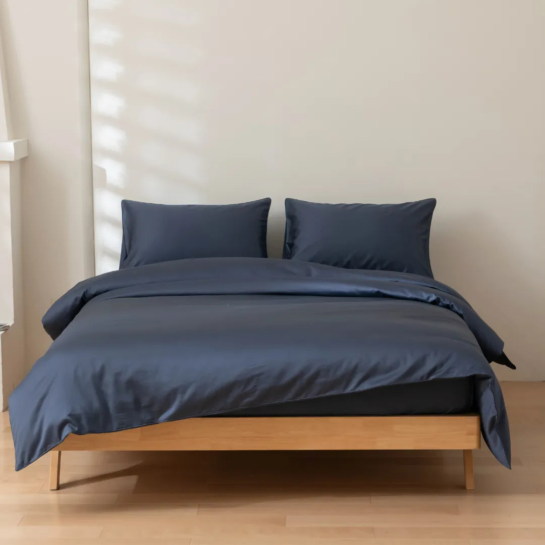 A neatly made bed with a Luxe Sateen Quilt Cover in Midnight from Linenly and two matching pillows on a simple wooden bed frame in a room with soft natural lighting.