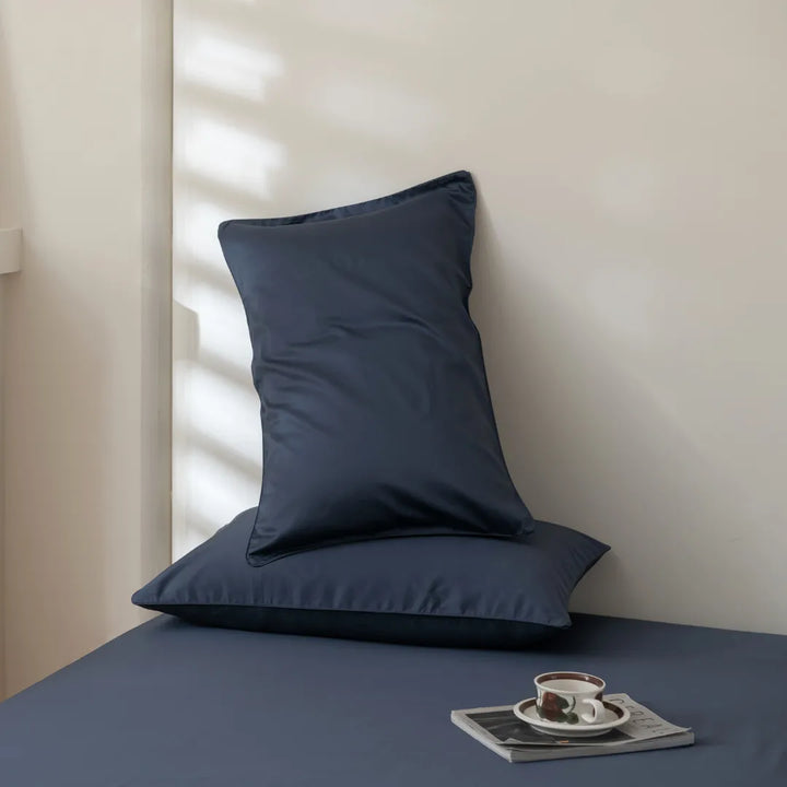 A neatly arranged dark blue pillow leans against another one on a matching bedspread made of Linenly Luxe Sateen Sheet Set - Midnight, and a book and a cup of coffee on a