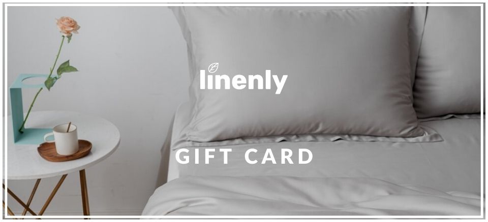 A neatly made bed with gray sheets and pillows is in the background. A small table holds a pink rose in a vase and a cup. The text "Linenly Gift Card" is centered over the image.
