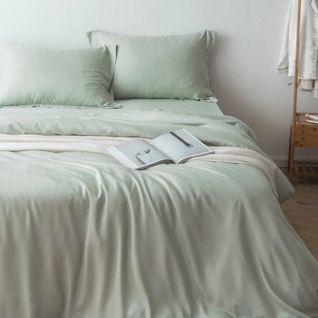 A woman in a nightgown is making a bed, gracefully flipping a white sheet. The room is softly lit, featuring framed artwork on the wall, a white clock, and neutral-colored decor.