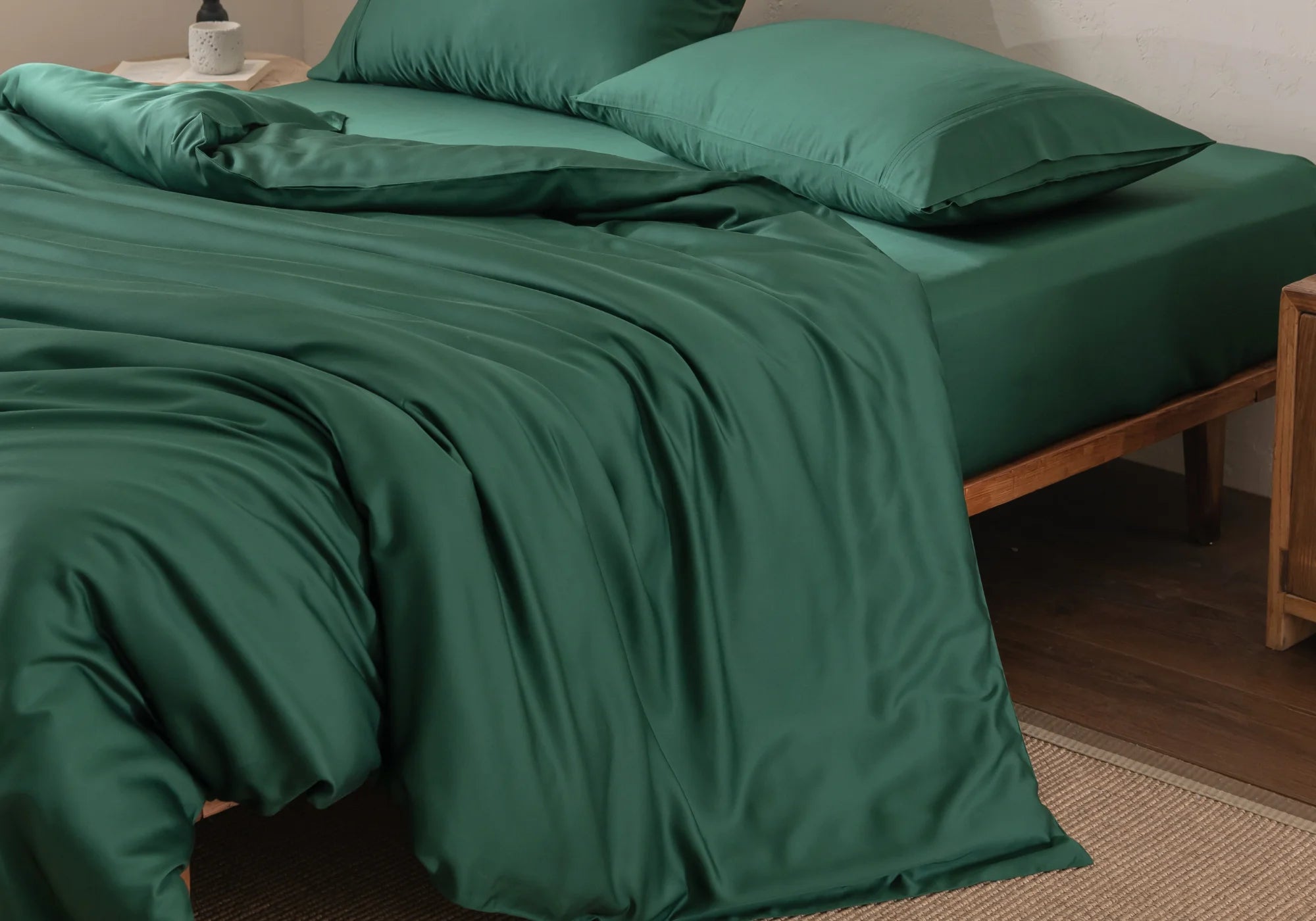 A neatly made bed with deep green, silky sheets and pillowcases. A wooden nightstand with a small lamp and bottle sits beside the bed on a brown rug and wooden floor.