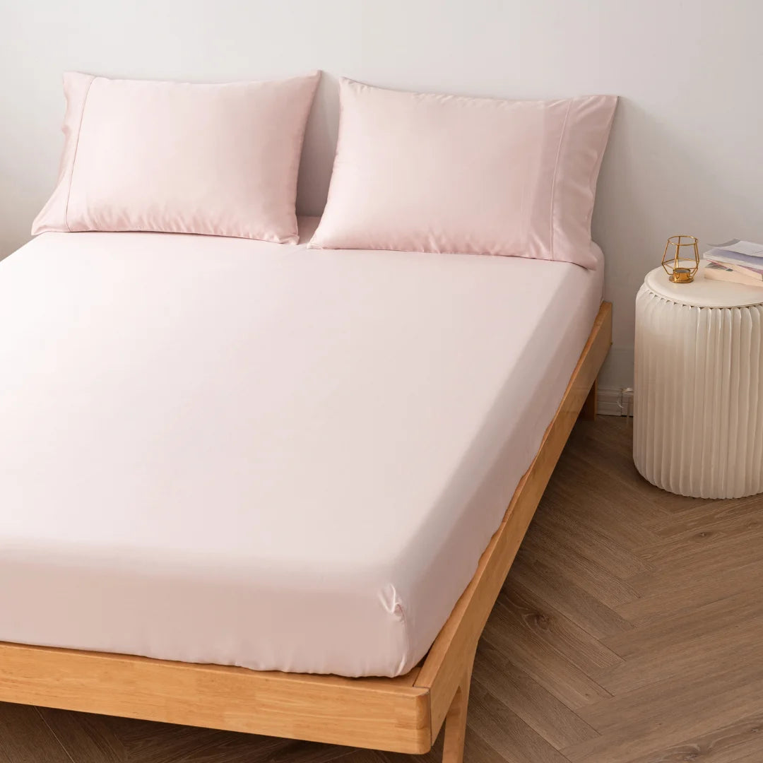 A neatly made bed with Linenly's Blush Bamboo Fitted Sheet and matching pillows in a bedroom with wooden flooring and a small side table.