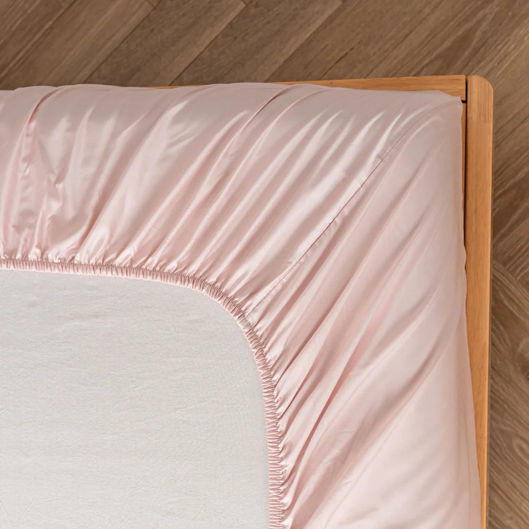 A close-up of a neatly made bed corner with a wooden bed frame, showcasing smooth Linenly Blush Bamboo Fitted Sheets made from sustainable bamboo fabric on a mattress.