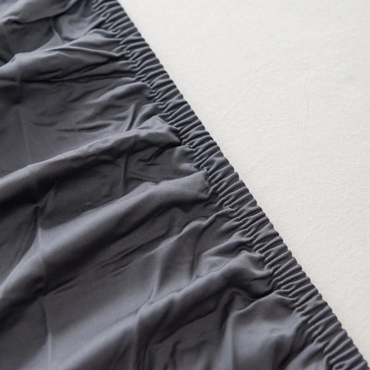 A close-up view of a section of Linenly's Bamboo Fitted Sheet in Charcoal, with an elastic edge on a pale, textured background.