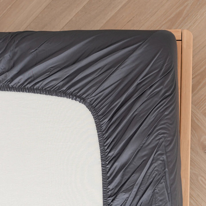 A fitted Linenly Charcoal Bamboo Fitted Sheet neatly covers a corner of a mattress on a wooden bed frame, showcasing a tidy bedroom setup.