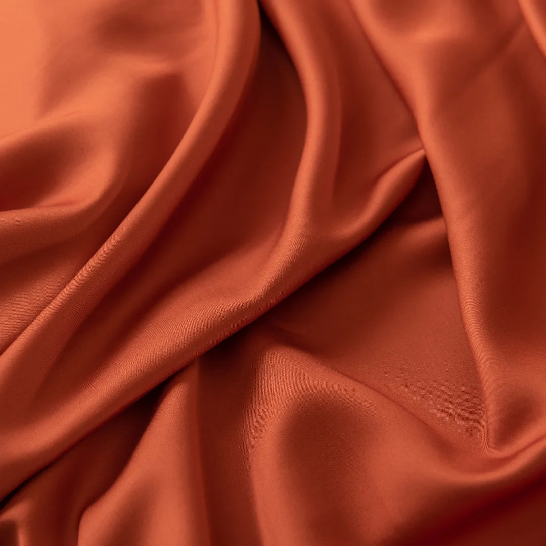 The Linenly Bamboo Fitted Sheet in Copper Sunset is captured close-up, displaying its smooth texture and gentle folds. Its rich hue and luxurious sheen exude sophistication and comfort, reminiscent of rust-colored silk fabric.