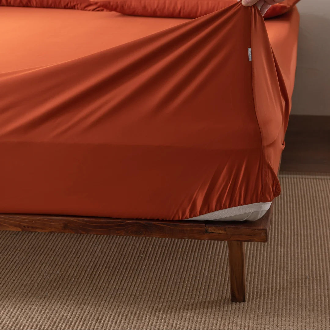 Someone is placing the Linenly Bamboo Fitted Sheet in Copper Sunset onto a mattress. A wooden bed frame rests on a beige woven rug as they carefully tuck in the sheet's corner for comfort.