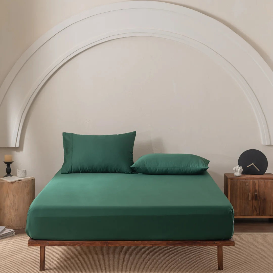 A minimalist bedroom features a wooden bed frame with a Linenly Bamboo Fitted Sheet Set in Evergreen and a green bedspread. It includes a sculptural white arch, wooden nightstand with decor, wooden block with candle, and is elegantly covered by a light-colored rug.