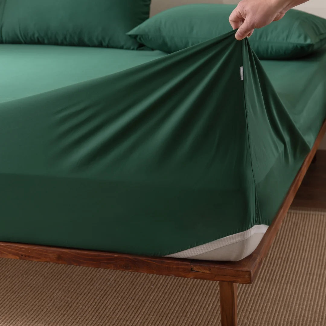 A hand gently lifts the Linenly Bamboo Fitted Sheet - Evergreen on a wooden bed frame. The deep green fabric, with matching pillowcases, exudes softness. Beneath it, a beige woven rug enhances the peaceful atmosphere.