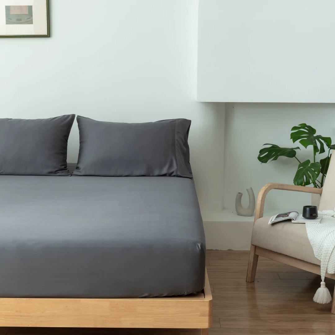 A minimalist bedroom with a neatly made bed featuring Linenly's granite grey bamboo fitted sheet with a deep-pocket design, accompanied by a wooden side table with a plant, a book, and a cup on it.
