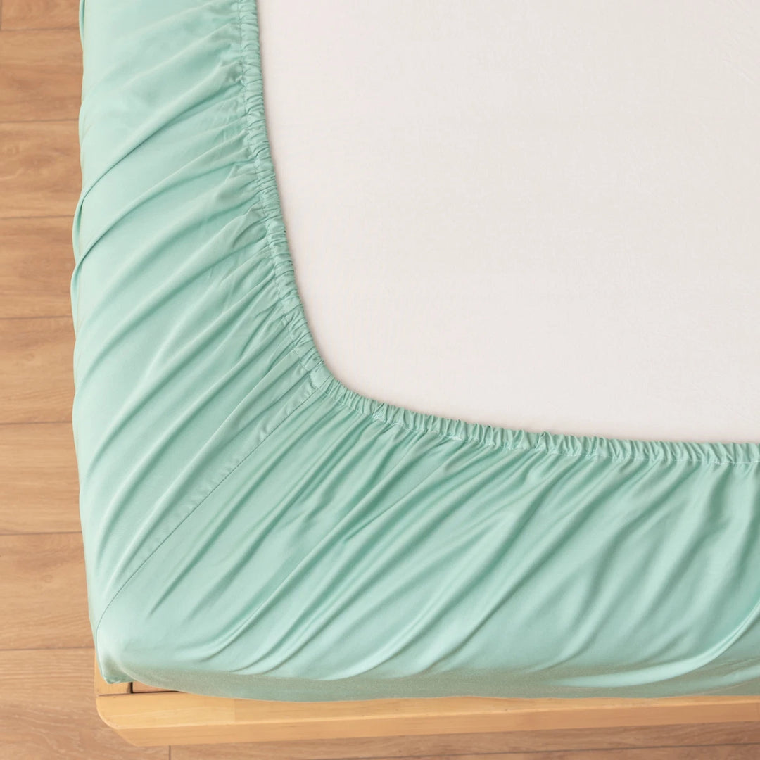 A corner of a neatly made bed with sustainable luxury bedding, including Linenly's Bamboo Fitted Sheet - Green Sheen on a mattress atop a wooden bed frame, showcasing the tight and crease-free elasticated corners.