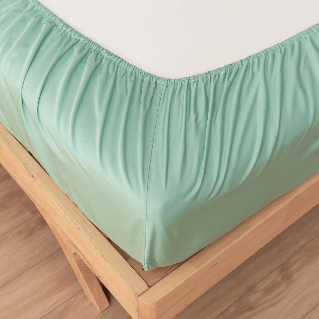 A Green Sheen Bamboo Fitted Sheet, made from breathable bamboo fabric, neatly wrapped around a mattress on a wooden bed frame, showcasing a clean and orderly bedroom setting by Linenly.