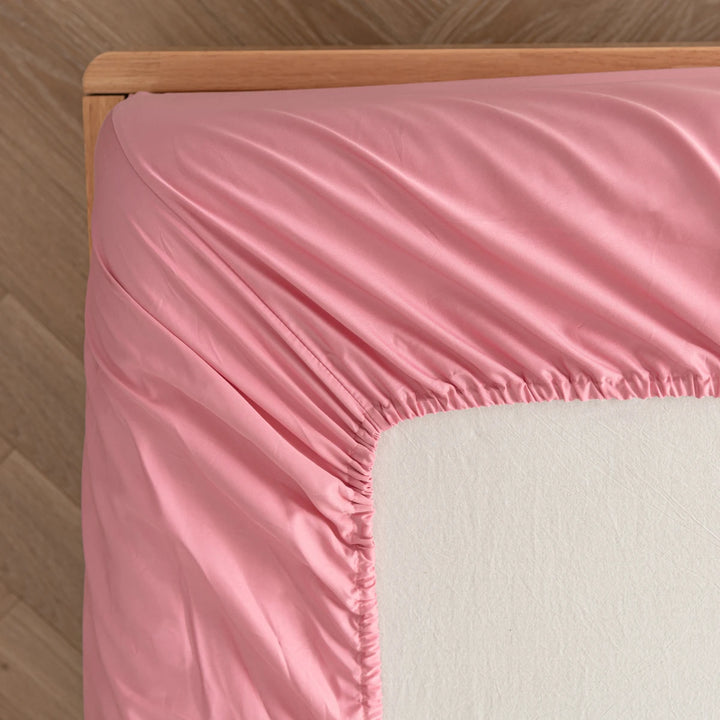 A neatly made bed with a smooth Linenly Bamboo Fitted Sheet in Light Rose and a comfortable beige mattress visible at the corner.
